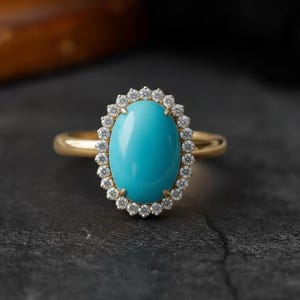 Könnte beinhalten: Ein goldener Ring mit einem ovalen türkisfarbenen Edelstein, umgeben von einem Kranz aus kleinen, klaren Steinen. Der Ring liegt auf einer dunklen, strukturierten Oberfläche, die das leuchtende Blau des Mittelsteins hervorhebt.