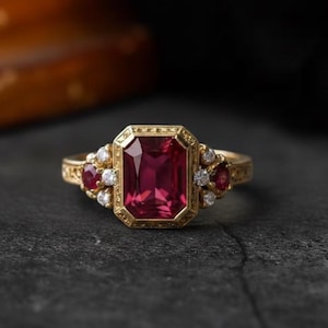 May include: A gold ring with a large, rectangular, deep red gemstone as the centerpiece. The ring is further adorned with smaller red and white gemstones, set in an ornate, vintage-style design. The ring is displayed on a dark, textured surface.