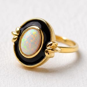 Könnte beinhalten: Ein goldener Ring mit einem ovalen, irisierenden Opal-Cabochon, der in einen glatten schwarzen Emaille-Rand eingefasst ist. Das Ringband besteht aus poliertem Gold und die Fassung hat kleine dekorative blattartige Goldakzente an den Seiten.