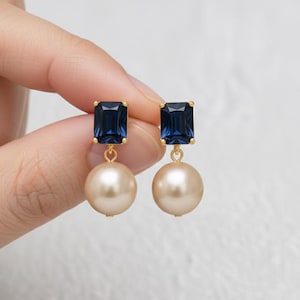 Puede incluir: Un par de pendientes colgantes con una piedra preciosa azul oscuro rectangular de corte esmeralda engastada en oro, suspendida sobre una perla redonda de color crema. Los pendientes se muestran juntos, destacando los engastes de metal dorado y el contraste entre las piedras azules y las perlas esféricas lisas.