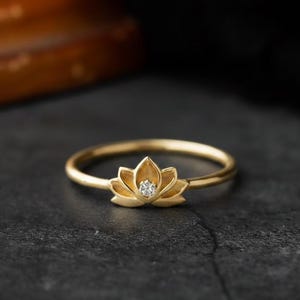 Puede incluir: Un anillo dorado con un diseño de flor de loto. La flor tiene pétalos detallados y una pequeña gema transparente en el centro. El anillo está sobre una superficie oscura y texturizada.