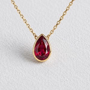 May include: A gold chain necklace featuring a single pear-shaped, deep red gemstone pendant set in a smooth gold bezel. The delicate metal chain connects to the top of the teardrop-shaped stone, which displays a faceted surface and rich colour.