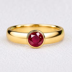 Puede incluir: Un anillo de oro amarillo pulido con un rubí redondo facetado de color rojo intenso engastado en un bisel liso. La banda es sólida y redondeada, mostrando un acabado metálico de alto brillo que contrasta con la vibrante piedra roja.