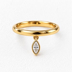 May include: A polished gold band ring featuring a single marquise-cut clear gemstone charm that hangs from a small circular loop. The ring has a smooth, rounded profile and a bright, reflective metallic finish, showcasing a simple and elegant design.