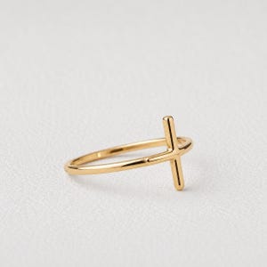 Könnte beinhalten: Ein polierter Goldring mit einer dünnen, glatten Ringschiene, die oben in eine vertikale Kreuzform übergeht. Das Metall hat eine reflektierende, hochglänzende Oberfläche und das Design besteht aus einer einfachen Linie, die das Kreuz bildet.