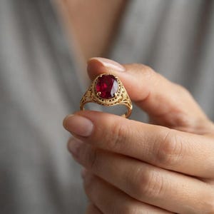 Pode incluir: Um anel de ouro com uma pedra preciosa de cor vermelho-escura de corte oval, montada numa banda de metal com filigrana decorativa. O engaste inclui um trabalho de metal intrincado que rodeia a pedra central de cor carmesim saturada.