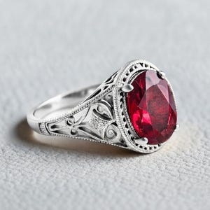 Pode incluir: Um anel de tom prateado com uma grande pedra preciosa vermelho-escura de corte oval, engastada numa moldura de metal decorativa. A banda apresenta detalhes intrincados de filigrana aberta nas laterais e uma borda perlada ao redor da pedra.
