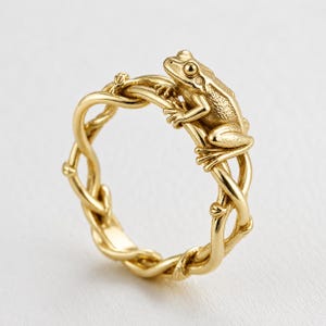 Puede incluir: Un anillo de oro con una rana detallada posada sobre una banda diseñada para parecerse a enredaderas entrelazadas. El metal tiene un acabado pulido y la rana está colocada en la parte superior de la banda, mostrando piel texturizada.