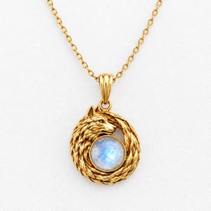 May include: A gold-toned necklace featuring a circular pendant with a sculpted wolf head design. The wolf is depicted in profile, encircling a round, iridescent moonstone cabochon. The pendant hangs from a delicate gold-toned link chain.