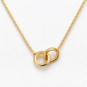 May include: A gold-toned necklace featuring a delicate chain and a pendant composed of two interlocking circular rings. The metal has a polished, reflective finish, and the design is centred on the chain, creating a symmetrical appearance.