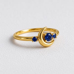 Puede incluir: Un anillo de oro con forma de media luna y tres piedras redondas de color azul intenso. La piedra central está engastada en la curva de la luna, mientras que dos piedras azules más pequeñas se sitúan a ambos lados de la fina banda de oro pulido.