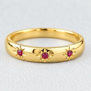 Peut inclure: Une bague en or poli ornée de trois pierres précieuses rondes d'un rouge éclatant, serties dans des motifs d'étoiles gravés. La surface métallique est lisse et réfléchissante, présentant une teinte dorée uniforme sur tout l'anneau.