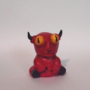 May include: A small, red, hand-sculpted figurine of a creature with two black horns, large yellow eyes with black pupils, and black feet. The creature is in a seated position with a smooth, rounded head and a simple, neutral facial expression.