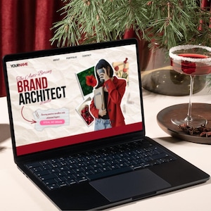 May include: A black laptop displaying a website with the text "BRAND ARCHITECT" and a photo of a person in a red blazer. A cocktail with a sugared rim sits nearby. The scene is set on a white surface with festive greenery.