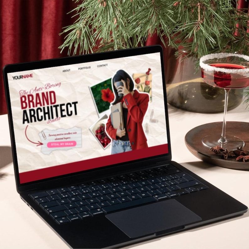 May include: A black laptop displaying a website with the text "BRAND ARCHITECT" and a photo of a person in a red blazer. A cocktail with a sugared rim sits nearby. The scene is set on a white surface with festive greenery.