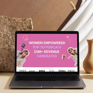 May include: A laptop displays a pink screen with the text "WOMEN EMPOWERED TOP 50 PODCAST $5M+ REVENUE GENERATED." The laptop is open on a light-colored surface, with a beige vase and a stone-like object in the background.
