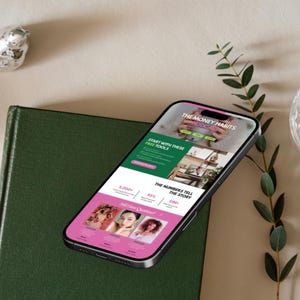 May include: A smartphone displays a webpage for "The Money Habits Podcast" with pink and green accents. The screen shows the text "Start with these free tools" and "The numbers tell the story". The phone rests on a green book.