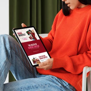 May include: A person holding a tablet displaying a website with the text "Brand Architect" and "The Death of Generic Content." The website features images and a red and white color scheme. The person is wearing an orange sweater and blue jeans.