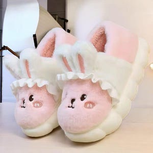 May include: A pair of plush bunny slippers. The slippers are primarily white and pink, with bunny faces on the toes and fluffy ears. The slippers have a cloud-like sole.