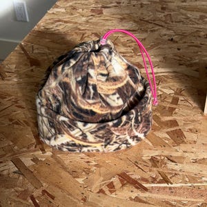 May include: A camouflage-patterned beanie hat with a drawstring closure and a bright pink cord. The hat is made of a soft, textured fabric and is designed to be worn on the head. The hat is sitting on a wooden surface.