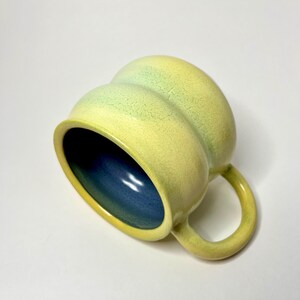 May include: A ceramic mug with a rounded, bulbous shape featuring a yellow and light green exterior with a visible crackle glaze texture. The interior of the mug is a deep, solid blue, and it includes a curved handle on the side.