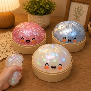 Könnte beinhalten: Drei runde, knödelartige Nachtlichter mit lächelnden Gesichtern, die jeweils auf einem beigen Sockel stehen. Eines ist rosa mit Glitzer, eines blau mit Glitzer und eines weiß mit irisierendem Finish. Ein kleines, klares, mit Glitzer gefülltes Spielzeug wird gehalten.