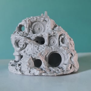 Puede incluir: Una miniatura de resina de color gris claro sin pintar que representa una vivienda en una ladera de varios niveles. La estructura presenta varias puertas circulares, entradas arqueadas y superficies rocosas texturizadas con escaleras.
