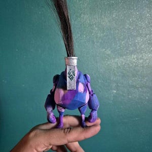 Puede incluir: Una pequeña figura impresa en tres dimensiones con un cuerpo redondeado con un patrón de camuflaje morado, azul y rosa. Tiene una pieza superior cilíndrica plateada con un largo mechón de pelo marrón oscuro que se extiende hacia arriba.