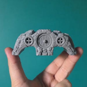 May include: A small, grey, unpainted 3D-printed miniature of a stone dwelling with a central circular door and two smaller circular windows on either side. The structure features a textured, rocky exterior surface with visible brick-like details.