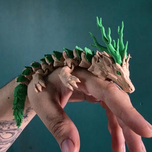 Puede incluir: Una figura de dragón articulada impresa en 3D con un cuerpo color canela, escamas texturizadas y detalles en verde vibrante a lo largo de la columna, la cabeza y la cola. La criatura tiene una forma serpentina con articulaciones visibles.