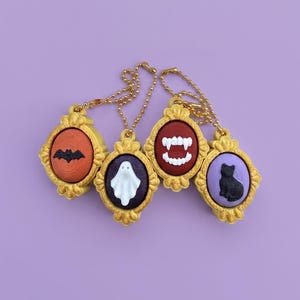 May include: Four oval-shaped gold-colored pendants with ball chains. Each pendant features a raised icon: a black bat on orange, a white ghost on purple, white vampire fangs on red, and a black cat on light purple, all set in gold frames.