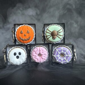 May include: Five square keychains featuring Halloween-themed circular charms. The charms include an orange jack-o'-lantern, a light green spider, a white ghost with black eyes and mouth, a pink brain, and a purple donut with a white spiderweb design.