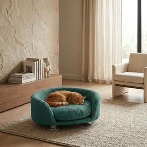 Op de afbeelding: Een tealkleurig huisdierbed met een rond ontwerp en chromen poten. Een oranje tabbykat slaapt in het bed. Het bed is gemaakt van een zacht, getextureerd materiaal.