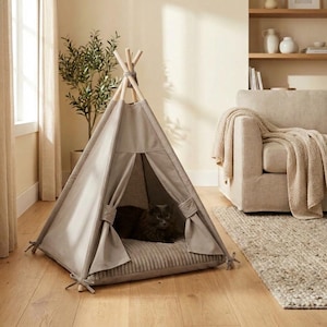 Puede incluir: Un tipi gris para mascotas con postes de madera y una atadura de cuerda en la parte superior. La tienda tiene una cama de estilo pana en el interior, y un gato negro descansa dentro.