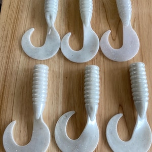 May include: Six white, soft plastic fishing lures with a curved tail and ribbed body. The lures are arranged on a wooden surface. The lures have a glittery appearance.