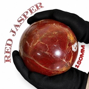 Op de afbeelding: Een grote, gepolijste rode jaspis bol met natuurlijke bruine en witte aders. De tekst RED JASPER staat in het rood boven de bol gedrukt en de tekst 150MM staat in het rood aan de rechterkant, wat een diameter van 150 millimeter aangeeft.