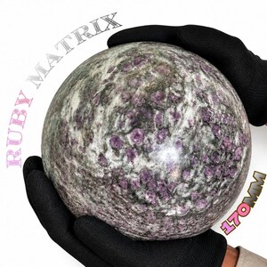 Könnte beinhalten: Ein großer, kugelförmiger Rubin-Matrix-Stein mit einer gesprenkelten Oberfläche, bei dem tief magentafarbene Rubineinschlüsse in eine graue und grüne Mineralbasis eingebettet sind. Der Text RUBY MATRIX und 170MM gibt die Größe des Steins an.