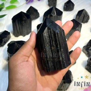 Könnte beinhalten: Eine Hand hält einen großen, schwarzen Turmalinkristall, umgeben von mehreren anderen schwarzen Turmalinkristallen unterschiedlicher Größe. Die Kristalle haben eine strukturierte Oberfläche und ein dunkles, glänzendes Aussehen. Der Hintergrund ist ein weiches, weißes Gewebe mit Andeutungen von Grün und Lila.