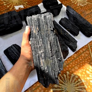 Puede incluir: Una colección de cristales de turmalina negra en bruto. Una mano sostiene una pieza grande, mostrando su textura estriada. Otras piezas de varios tamaños están dispuestas sobre una superficie blanca, con un fondo estampado.