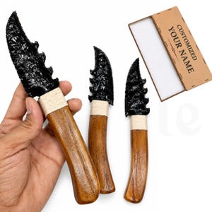 Puede incluir: Tres cuchillos con hojas de obsidiana negra dentada y mangos de madera marrón envueltos en cordel claro. Se incluye una caja de cartón rectangular con el texto CUSTOMIZED YOUR NAME impreso en la parte superior en letras mayúsculas negras.