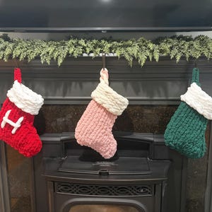 Könnte beinhalten: Drei grob gestrickte Weihnachtsstrümpfe hängen in einer Reihe. Der linke Strumpf ist leuchtend rot mit einem weißen Buchstaben H und einem weißen Umschlag. Der mittlere Strumpf ist hellrosa mit einem cremefarbenen Umschlag. Der rechte ist dunkelgrün.