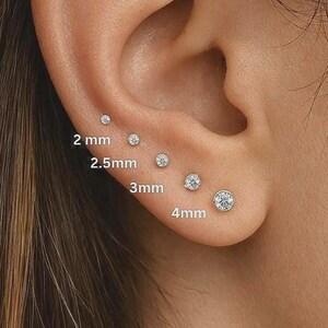 May include: A set of five round, clear stud earrings arranged in a row on an earlobe, with sizes labeled as 2 mm, 2.5 mm, 3 mm, and 4 mm. Each earring features a single, brilliant-cut stone set in a metallic bezel, showing a clear size progression.