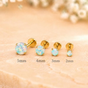 May include: Four gold-colored stud earrings with iridescent, light blue opal stones. The sizes are 5mm, 4mm, 3mm, and 2mm. The earrings are displayed on a light brown surface with a blurred floral background.