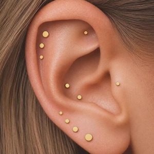 May include: A collection of ten small, circular, gold-toned stud earrings arranged along the cartilage, conch, and lobe of an ear. The studs vary slightly in size, featuring a smooth, flat, polished surface that reflects light consistently.