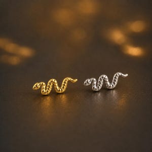 May include: Two small stud earrings shaped like snakes, one in a polished gold tone and the other in a polished silver tone. Each earring features a textured, winding body design with a defined head and a curved tail, presented as a pair of studs.