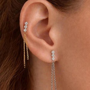 May include: Earrings with three small, clear gemstones in a vertical line, connected to a delicate chain. One earring has a gold chain, the other a silver chain. The earrings are worn on the ear.