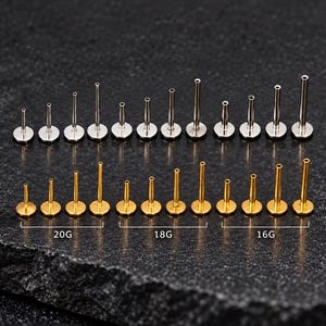 May include: A collection of silver-toned and gold-toned flat back labret studs arranged in rows. The studs vary in length and are categorized by gauge sizes labeled as 20G, 18G, and 16G. Each piece features a circular base and a hollow post.