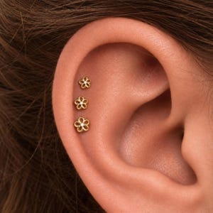May include: Three gold-toned flower-shaped stud earrings arranged vertically along the upper ear cartilage. Each earring features a delicate open-petal design with a small, clear, sparkling stone set in the center of the floral arrangement.