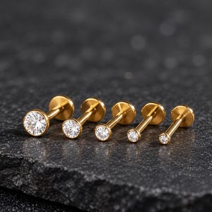 May include: Five gold-toned labret studs arranged in a row, each featuring a round, clear, faceted gemstone set in a bezel. The studs vary in size, with the largest on the left and the smallest on the right, each ending in a flat circular back.