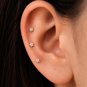 May include: Three small gold stud earrings shaped like flowers, each featuring a central gold bead surrounded by six tiny clear stones. These delicate studs are arranged vertically along the outer edge of the ear cartilage and lobe.